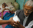 Older, thin, sickly woman lies on cot in background, older man at the foot of the cot receives container from someone standing