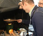 Man holds instrument to measure emissions over lit wood-burning stove, which burns under a hood, woman holds up hood