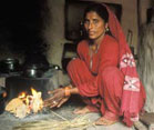 woman, barefoot, squats on the floor next to a cooking fire, dark smoke fills the air, no visible chimney or ventilation