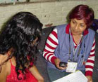 Two women sit on a sofa together, one faces the camera, holding a handheld device, the other's hair covers her face