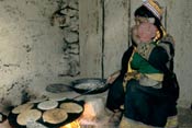 Woman squats, toddler on her lap, cooks large pan full of flat bread over indoor cookstove, flames visible beneath pan