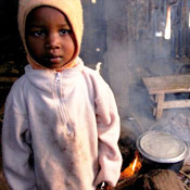 Young child in sweatshirt and hood looks into camera, fire burns with smoke in background