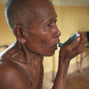 Older man uses inhaler, shirtless, bare floored clinical setting with empty bedframes in background