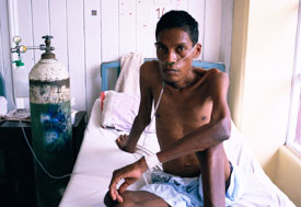 Emaciated man sits up in bed, shirtless, next to large tank with tubes in nose