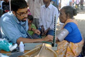 A male medical worker with stethoscope takes the blood pressure of an older woman, seated, in an outdoor clinic