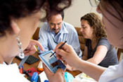 In foreground two medical workers view electronic device screen together, in background two more discuss together at a table