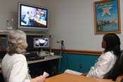 Two women seated at conference table, looking at teleconferencing equipment, with a screen that shows another conference room