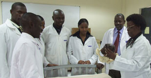 Group of medical students in white lab coats stands around hospital bed, woman leading discussion points to model she holds