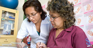 Two women lean over at desk to take notes, poster on pregnancy in background
