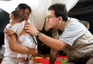 Doctor puts stethoscope on baby's back while mother holds the baby, all seated on ground, tarps in background