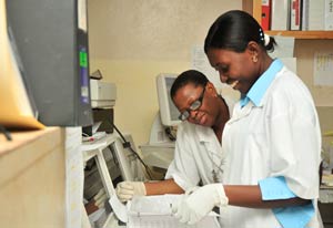 Two women in white lab coats wearing medical gloves review papers in three-ring binder