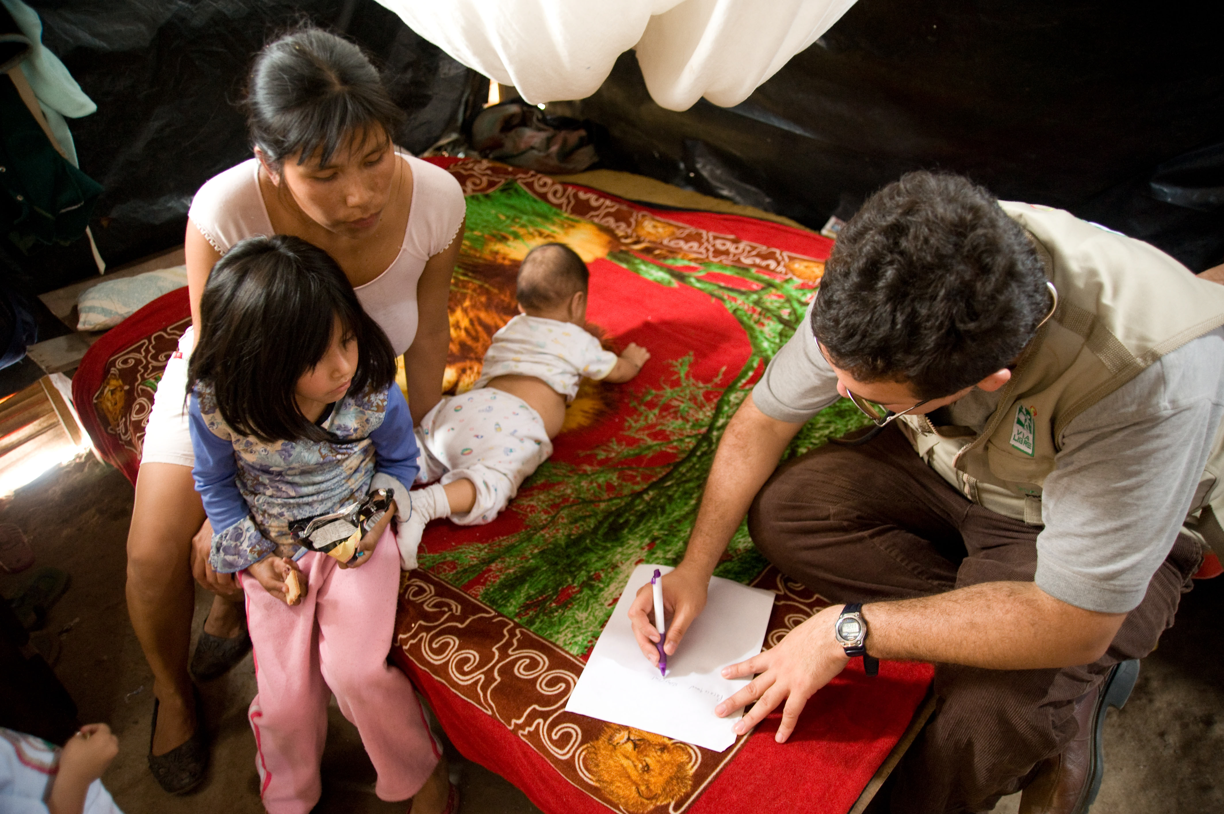 A researcher interviews a mother who is holding a young child while another child plays on the floor