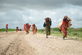 People carrying many large bundles walk along a dirt road.