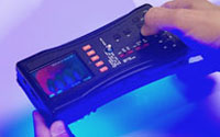 Photo courtesy of FDA, close up of hands holding FDA Counterfeit Detection Device-3 CD-3, black electronic box with many buttons emits glow on bottom