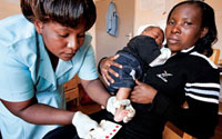 Photo by James Pursey/USAID, young woman holds a baby on her lap, a medical worker examines the baby, referencing an informational card
