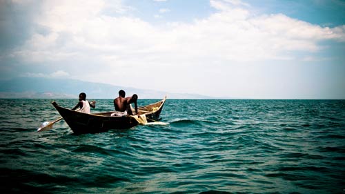 three young men in small wooden boat, fishing nets draped on side, wide expanse of blue water, mountains in distant background