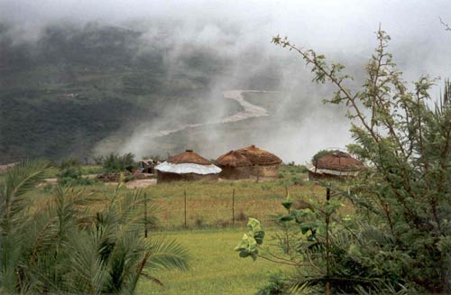 Group of four huts on hillside, mist rises above them, seen from higher on hill