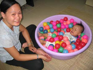 Baby lies in purple bucket full of balls, woman seated cross-legged on floor