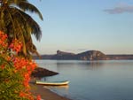 small, empty wooden boat rests on shore of beach, partially in water, red flowers and palm tree in foreground, cliffs and plains on other side of water