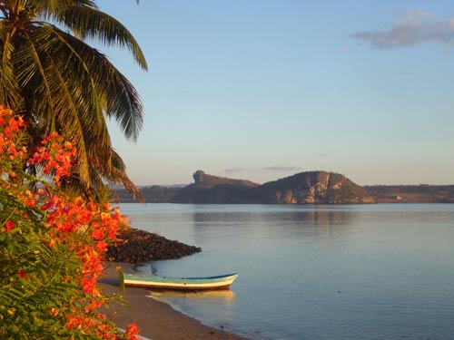 small, empty wooden boat rests on shore of beach, partially in water, red flowers and palm tree in foreground