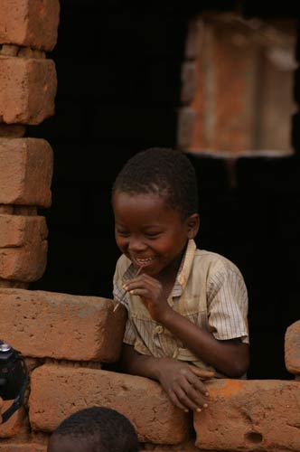 Smiling young Malawian boy leans out of bare window of rough brick building