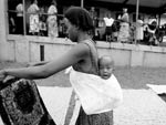 Young Ugandan woman, holding and looking at piece of fabric, with baby tied to her back, baby looks straight at camera