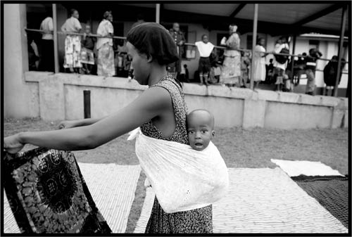 Young Ugandan woman, holding and looking at piece of fabric, with baby tied to her back, baby looks straight at camera 