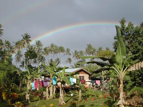 double rainbows in grey sky over house surrounded by palm trees and other greenery, laundry on line surrounds house