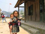 Young Vietnamese girl with boy toddler strapped to her back, standing in front of wooden house on dry dirt ground