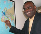 Dr. Clement Adebamowo looks at camera, smiling, points to Nigeria on a world map