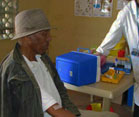 Older man seated in clinic holds out arm, bandage in crook of elbow, clinician in background arranges vials on table