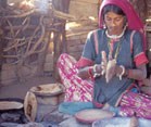 Woman sits cross-legged on ground rolling dough in hands, round flat bread cooking in pan on indoor cookstove