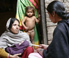 Woman holds swaddled baby in her arms, listening to woman across from her who has back to camera, another young child looks on