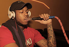 Young man exhales smoke after inhaling from hose of a waterpipe, wearing large headphones and baseball cap.