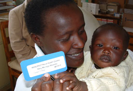 A Kenyan woman holding a baby holds up a blue and white patient ID card
