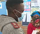 Researcher wearing a mask takes notes on a clipboard while interviewing a masked subject in a clinic.