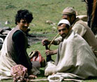 Indian men sit on the grass in a field, one smokes a water pipe