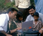 Outdoors, child seated at laptop is instructed by man in shirt and tie, adult and another child look on close by