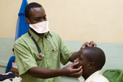 Photo: male doctor wearing a facemask, standing, examines a male patient, seated on hospital bed; bed net hang from walls