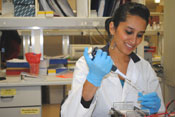 Photo: female researcher in white lab coat and blue latex gloves, smiling, lab in background
