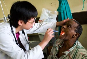 Woman doctor in white coat wears stethoscope, holds male patient's head back, uses pen light to look in his mouth