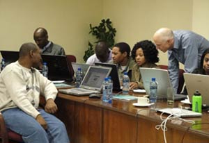 Seven people seated in front of many laptop computers on opposite sides of a long desk, one many looks over a woman’s shoulder