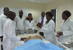 Group of medical students in white lab coats stands around hospital bed, woman leading discussion points to model she holds