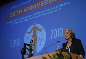 A woman at a podium speaks into a microphone, slide displays in background, reading 20th anniversary 1990 2010