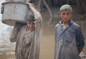 Two young men, mud on their shirts, stand in front of murky brown water, one holds large bucket on his shoulder