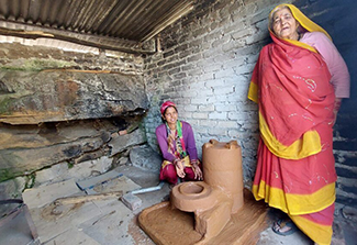 This photo shows two women, one is squatting behind next to a smokeless mud cookstove, the other is standing in front of the stove