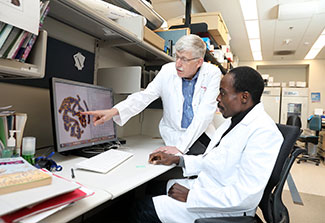 Picture of Dr. Francis Collins, standing, and Idowu Aimola, sitting. Both are wearing white lab coats and looking at a computer screen.