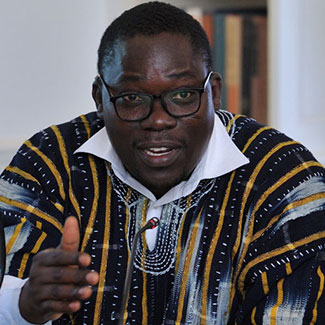 A photo of Dr. Thomas Hormenu wearing black-rimmed glasses, and a white-collared shirt under a black-and-gold, dashiki-type shirt.