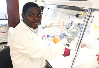 Picture of Dr. Kolapo Oyebola wearing a white lab coat and protective gloves, working with samples in a lab.