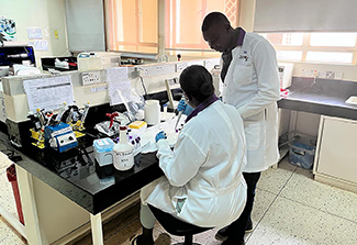 The photo on this page shows Dr. Alex Kayongo, a Ugandan man wearing a lab coat, consulting with a woman, similarly dressed in a lab coat. She sits and he stands at a desk in his laboratory